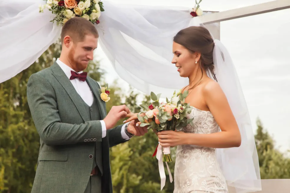 Ženích navlieka neveste svadobnú obrúčku počas romantického svadobného obradu pod kvetinovou výzdobou v exteriéri