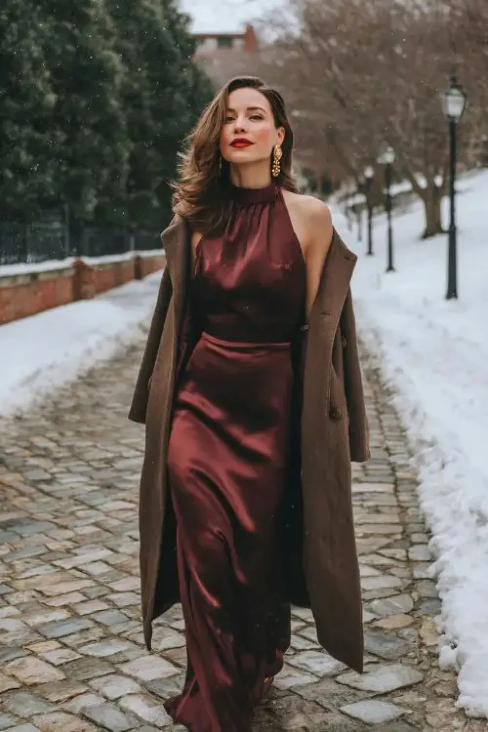 Elegantný outfit na zimnú svadbu – dlhé bordové saténové šaty s odhalenými ramenami doplnené hnedým kabátom a zlatými náušnicami